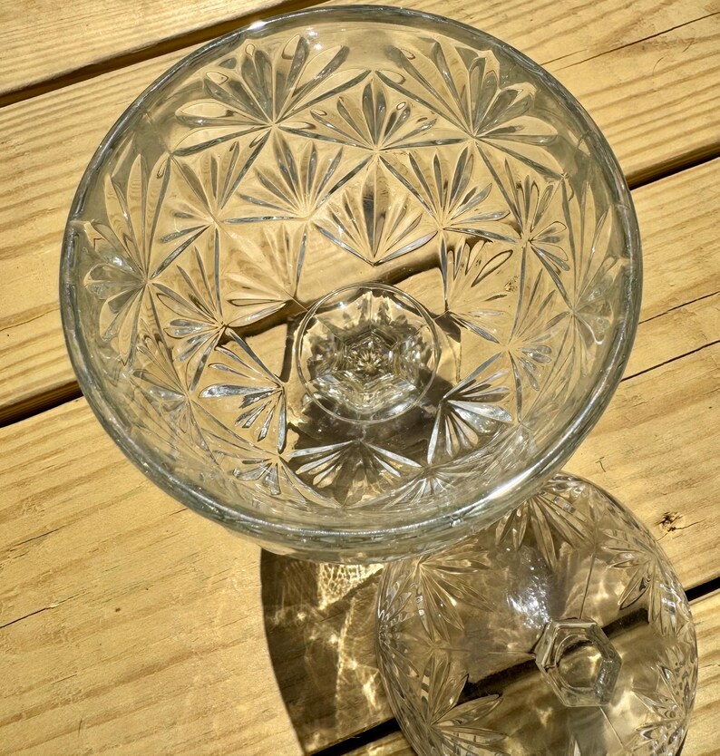 May include: Clear glass serving bowl with a pedestal base and a matching lid. The bowl and lid feature a detailed, repeating starburst pattern. The bowl is round and sits on a short, hexagonal base. The lid is also round with a hexagonal handle.