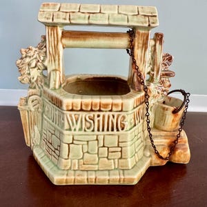 May include: A ceramic wishing well planter featuring a light green and brown stone-like finish. The structure includes a roof, a bucket suspended by a thin metal chain, and the embossed text WISHING WELL on the front. It has a hollow center.