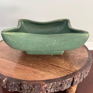 May include: A matte sage green ceramic bowl with a square base and a distinctively curved, undulating rim. The bowl features a smooth, solid-colored finish with a small, light-colored scratch visible on the front exterior surface of the vessel.