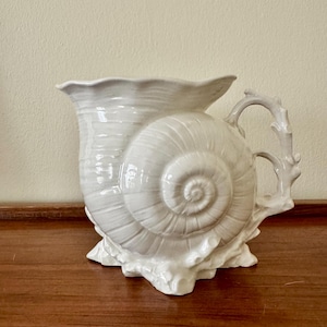May include: A white ceramic pitcher shaped like a large spiral seashell. The vessel features a textured, ridged surface mimicking a natural shell, a wide flared rim, and a handle designed to resemble a piece of coral with branching segments.