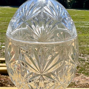 May include: A clear, cut-glass candy dish with a lid. The dish has a round, bulbous body and a faceted, hexagonal base. The lid is also cut glass and has a small, rounded handle. The dish is sitting on a wooden surface.