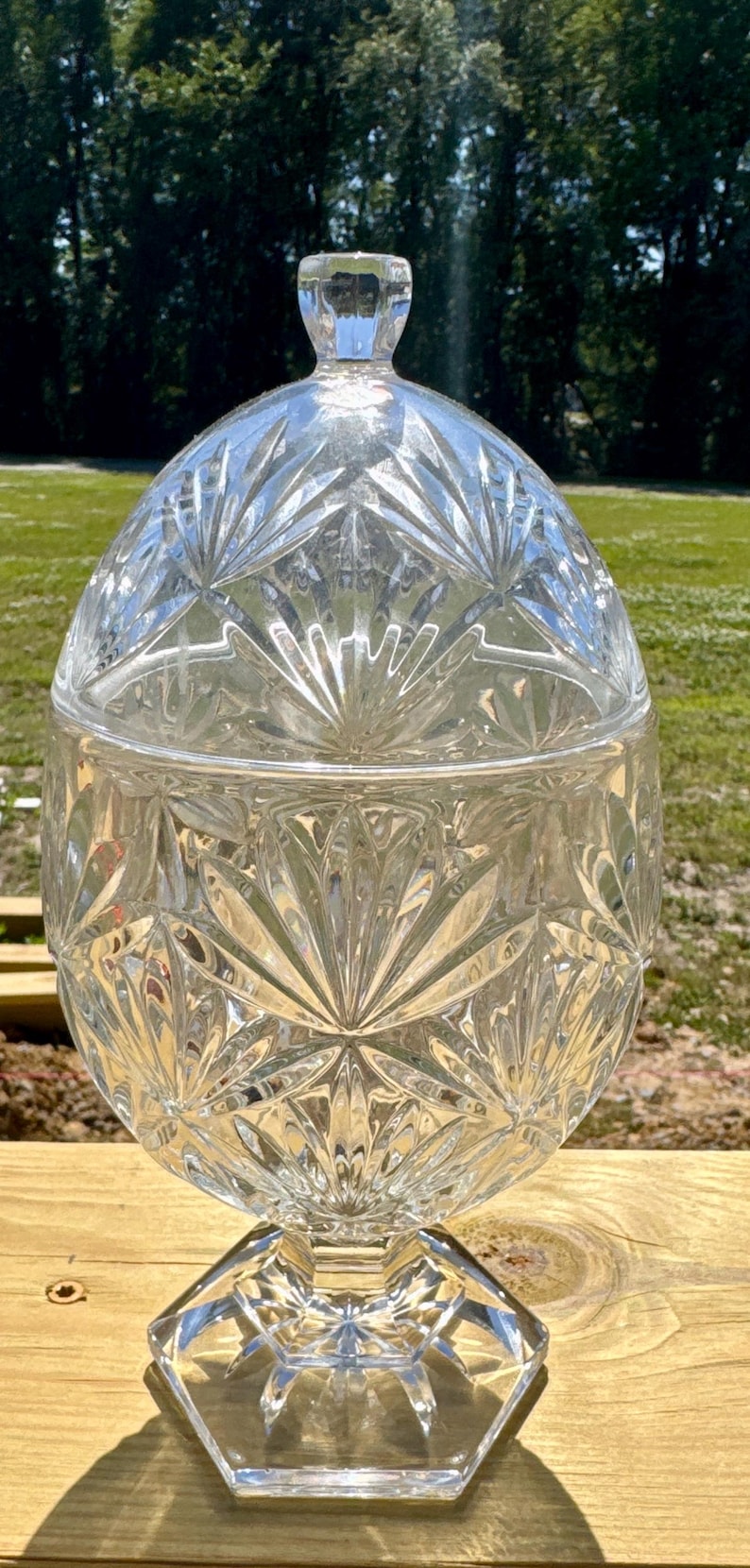 May include: A clear, cut-glass candy dish with a lid. The dish has a round, bulbous body and a faceted, hexagonal base. The lid is also cut glass and has a small, rounded handle. The dish is sitting on a wooden surface.
