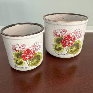 May include: Two cylindrical ceramic planters with a speckled off-white finish and a dark brown rim. Each planter features a hand-painted illustration of pink and red geranium flowers with green leaves and the word Geranium written in cursive script.