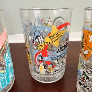 Op de afbeelding: Een helder drinkglas met kleurrijke Disney-personageafbeeldingen, waaronder Donald Duck, Sneeuwwitje en de zeven dwergen. Het glas bevat de tekst: "DONALD's middle name is Fauntleroy" en "Disney Stars & Motor Cars features classic animation from 1920 to 1990."