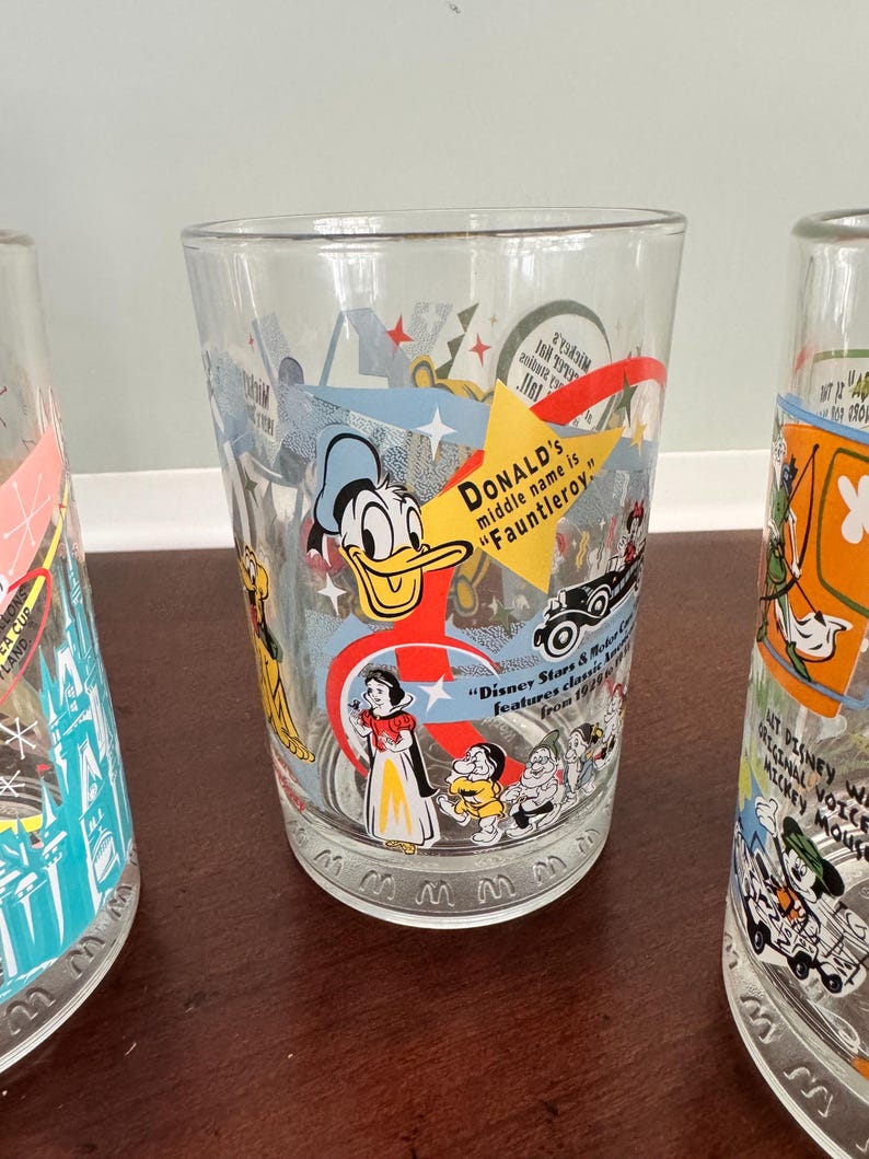 Op de afbeelding: Een helder drinkglas met kleurrijke Disney-personageafbeeldingen, waaronder Donald Duck, Sneeuwwitje en de zeven dwergen. Het glas bevat de tekst: "DONALD's middle name is Fauntleroy" en "Disney Stars & Motor Cars features classic animation from 1920 to 1990."