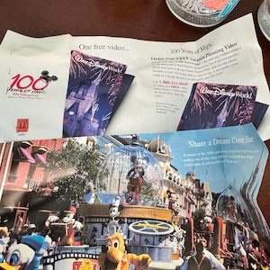 Op de afbeelding: Een promotionele papieren flyer voor de 100 Years of Magic viering van Walt Disney World. De flyer bevat de tekst One free video, 100 Years of Magic en Share a Dream Come True, naast kleurrijke afbeeldingen van Mickey Mouse en vrienden.