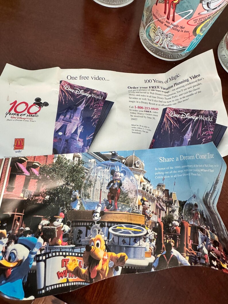 Op de afbeelding: Een promotionele papieren flyer voor de 100 Years of Magic viering van Walt Disney World. De flyer bevat de tekst One free video, 100 Years of Magic en Share a Dream Come True, naast kleurrijke afbeeldingen van Mickey Mouse en vrienden.