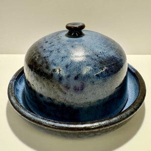 May include: A ceramic butter dish with a dome-shaped lid and a matching circular base. The piece features a mottled glaze in shades of deep blue, light blue, and black, with a smooth, glossy finish and a small knob handle on the lid top.