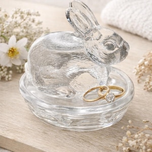 May include: A clear glass bunny-shaped ring holder with two gold rings. The bunny sits atop a small dish, perfect for storing jewelry. The design is detailed, with visible textures and a charming aesthetic.