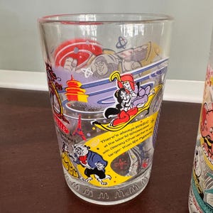 Op de afbeelding: Een helder drinkglas met kleurrijke illustraties van Disney-figuren, waaronder Buzz Lightyear, Jasmine, Aladdin, Belle en het Beest. Het glas bevat de tekst: There's always MAGIC at the World Showcase with Disney Characters and larger than life Puppets.