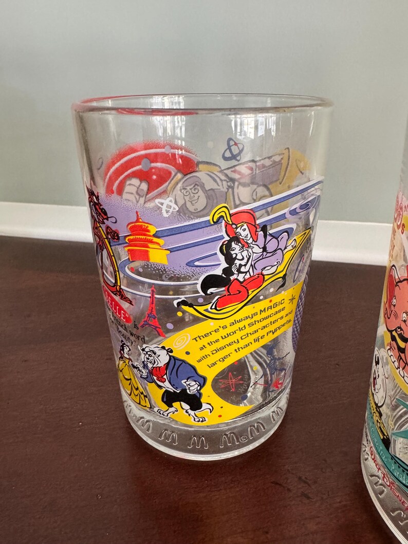 Op de afbeelding: Een helder drinkglas met kleurrijke illustraties van Disney-figuren, waaronder Buzz Lightyear, Jasmine, Aladdin, Belle en het Beest. Het glas bevat de tekst: There's always MAGIC at the World Showcase with Disney Characters and larger than life Puppets.