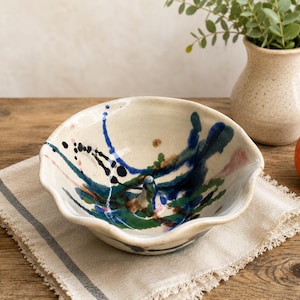 May include: A handmade ceramic bowl with a unique, abstract design. The bowl features a cream-colored base with splashes of blue, green, and pink paint. It sits on a linen napkin with a striped border, alongside a small vase and a red apple.