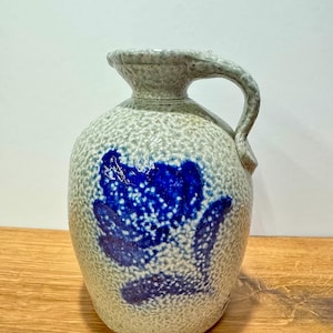 May include: A ceramic jug with a textured, speckled light gray finish and a single handle. The front of the jug features a prominent, hand-painted floral design in a deep, vibrant blue glaze, showcasing a stylized flower with leaves.