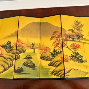May include: A folding screen with a Japanese landscape painting. The artwork features a yellow background with a mountain, river, bridge, and traditional houses. The painting uses shades of green, brown, red, and blue.