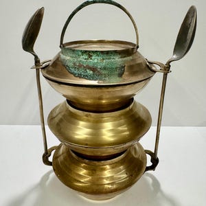 May include: A three-tiered brass food carrier featuring a rounded base and stacked bowls. The top section displays visible green oxidation on the metal surface. Two long metal spoons are attached to the sides, extending upward from the frame.