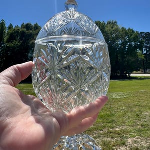 May include: A clear, cut-glass candy dish with a lid, held in a hand against a sunny, blue sky. The dish features a detailed, starburst pattern and sits on a pedestal base. The lid has a small, faceted knob.