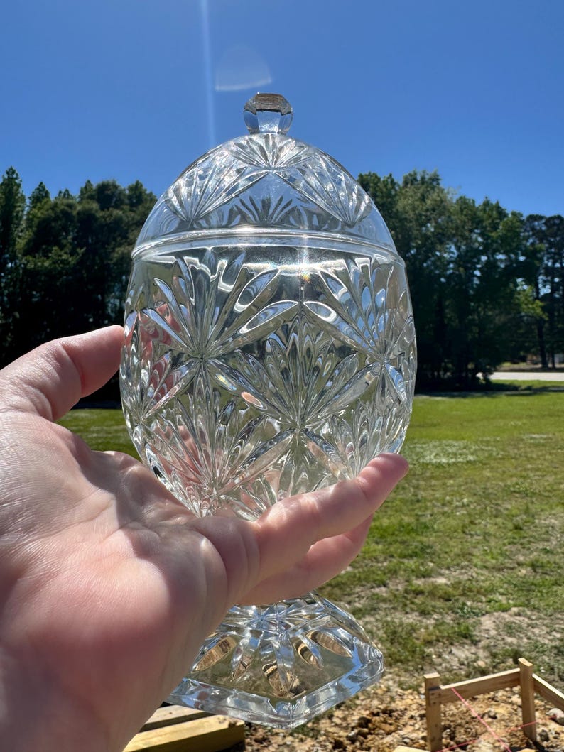 May include: A clear, cut-glass candy dish with a lid, held in a hand against a sunny, blue sky. The dish features a detailed, starburst pattern and sits on a pedestal base. The lid has a small, faceted knob.
