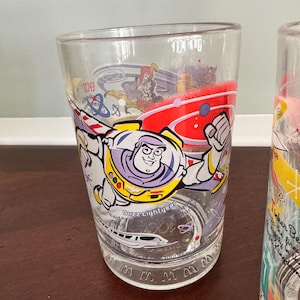 Op de afbeelding: Een helder drinkglas met een kleurrijke afbeelding van Buzz Lightyear in een ruimtepak met uitgestrekte vleugels. Het ontwerp bevat rode, gele en paarse accenten, een monorail-illustratie en de tekst Buzz Lightyear op het oppervlak.