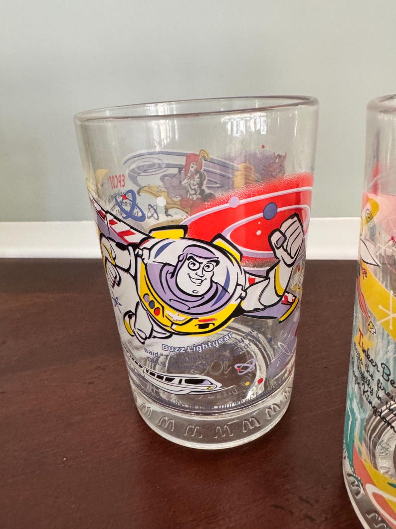 Op de afbeelding: Een helder drinkglas met een kleurrijke afbeelding van Buzz Lightyear in een ruimtepak met uitgestrekte vleugels. Het ontwerp bevat rode, gele en paarse accenten, een monorail-illustratie en de tekst Buzz Lightyear op het oppervlak.