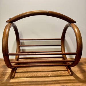 May include: A brown wooden magazine rack featuring a curved handle at the top, two large circular side frames, and a series of horizontal slats forming the base and sides to hold items. The structure is made of smooth, polished dark wood.
