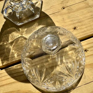 May include: Clear glass serving dish with a lid, featuring a starburst pattern and a faceted base. The lid has a hexagonal knob. The dish sits on a wooden surface, with sunlight casting shadows.