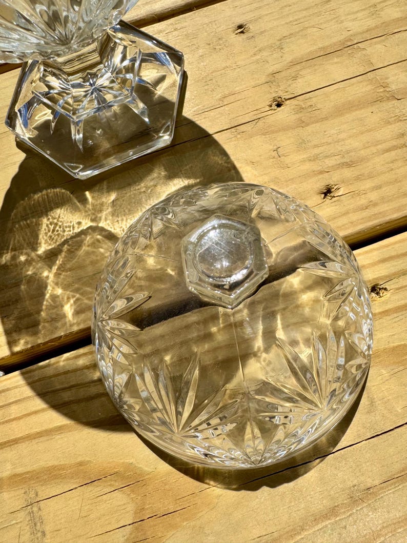 May include: Clear glass serving dish with a lid, featuring a starburst pattern and a faceted base. The lid has a hexagonal knob. The dish sits on a wooden surface, with sunlight casting shadows.
