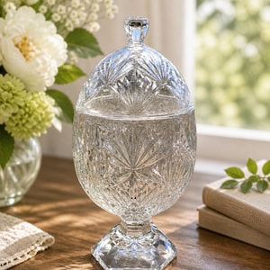May include: A clear, cut-glass candy dish with a lid, set on a hexagonal base. The dish has a detailed, starburst pattern. The lid has a small, faceted knob. The dish is on a wooden surface, with flowers and books in the background.
