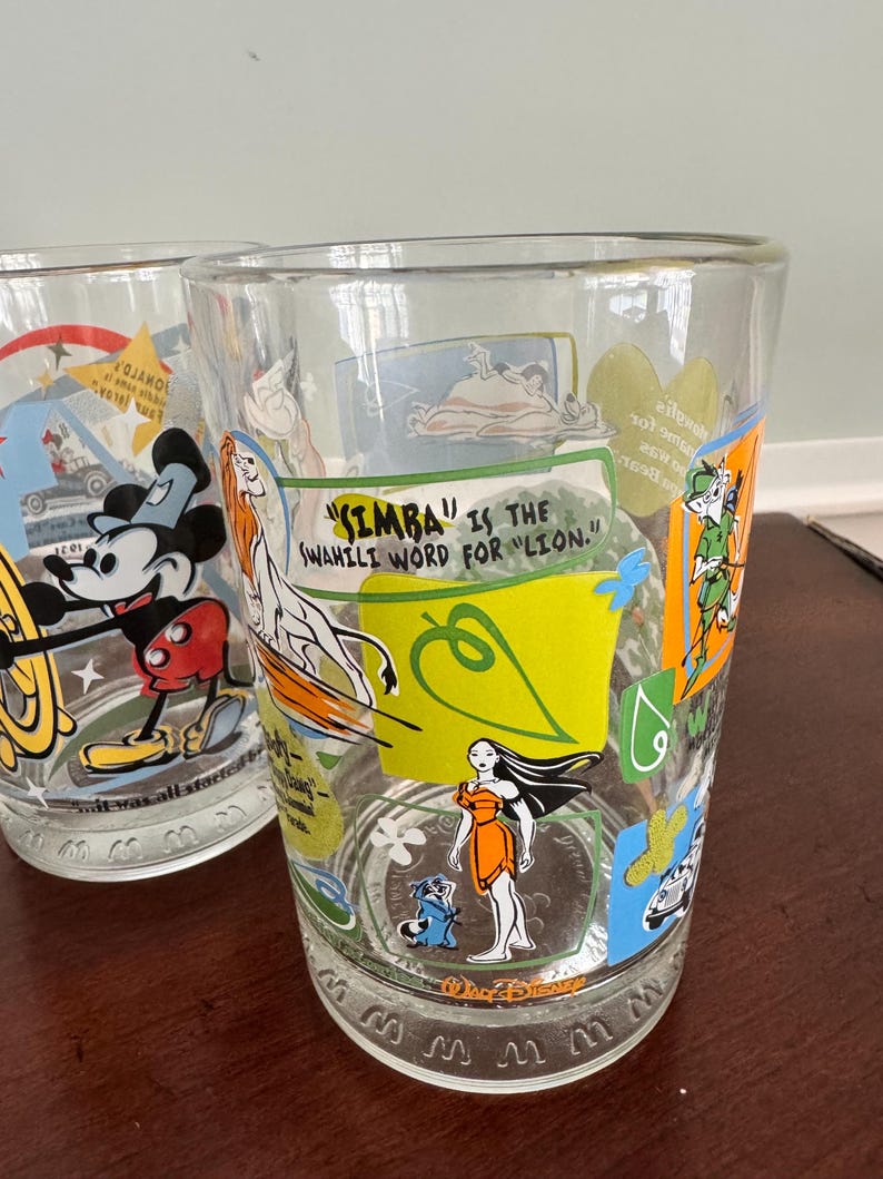 Op de afbeelding: Een helder drinkglas met kleurrijke Disney-personages zoals Pocahontas, Simba en Robin Hood. De tekst luidt: "Simba" is the Swahili word for "lion." De onderkant is voorzien van het Walt Disney-logo en een herhalend patroon.