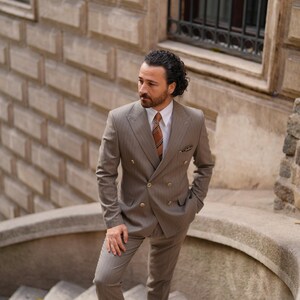 Puede incluir: Un traje marr&oacute;n claro de doble botonadura con un sutil patr&oacute;n de rayas finas. El traje incluye una chaqueta y un pantal&oacute;n a juego, combinados con una camisa blanca y una corbata estampada. La persona est&aacute; de pie en una escalera de piedra.