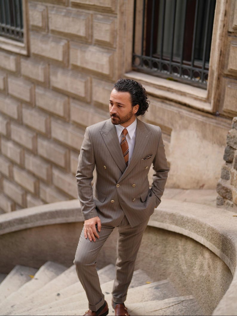 Puede incluir: Un traje marr&oacute;n claro de doble botonadura con un sutil patr&oacute;n de rayas finas. El traje incluye una chaqueta y un pantal&oacute;n a juego, combinados con una camisa blanca y una corbata estampada. La persona est&aacute; de pie en una escalera de piedra.