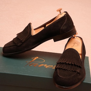 May include: Black suede loafers with fringed detailing. The shoes are presented on a teal box with gold lettering. The loafers have a classic design and are suitable for various occasions.