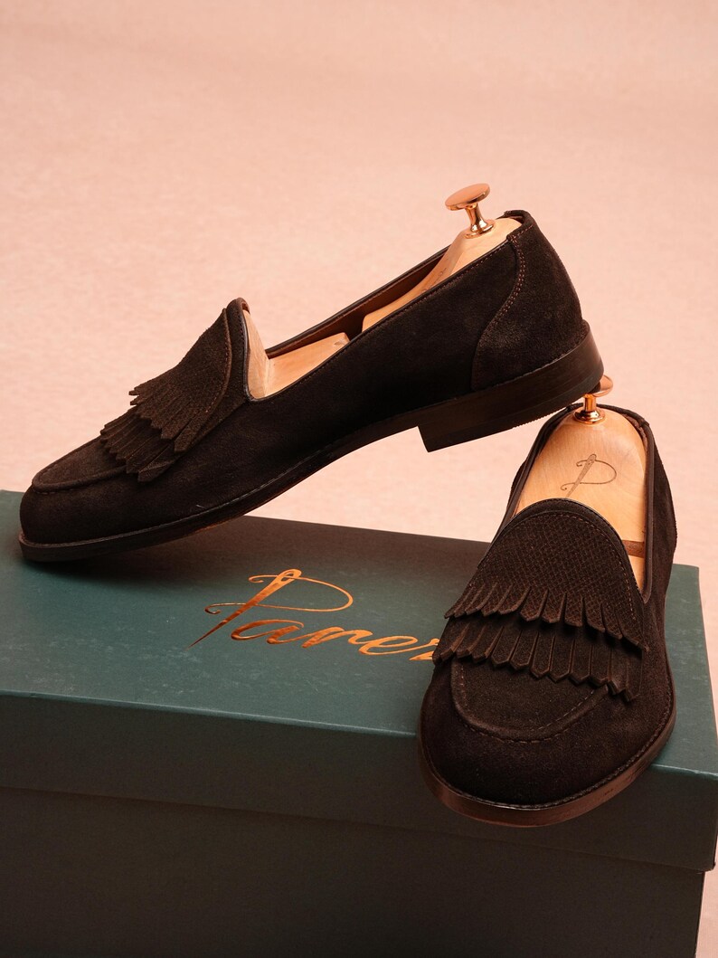 May include: Black suede loafers with fringed detailing. The shoes are presented on a teal box with gold lettering. The loafers have a classic design and are suitable for various occasions.
