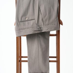 Puede incluir: Pantalones grises de raya diplom&aacute;tica con bolsillo abotonado y trabillas laterales ajustables, colgados en una percha de madera. Los pantalones tienen dobladillo con vuelta y un dise&ntilde;o cl&aacute;sico a medida, adecuado para vestimenta formal.