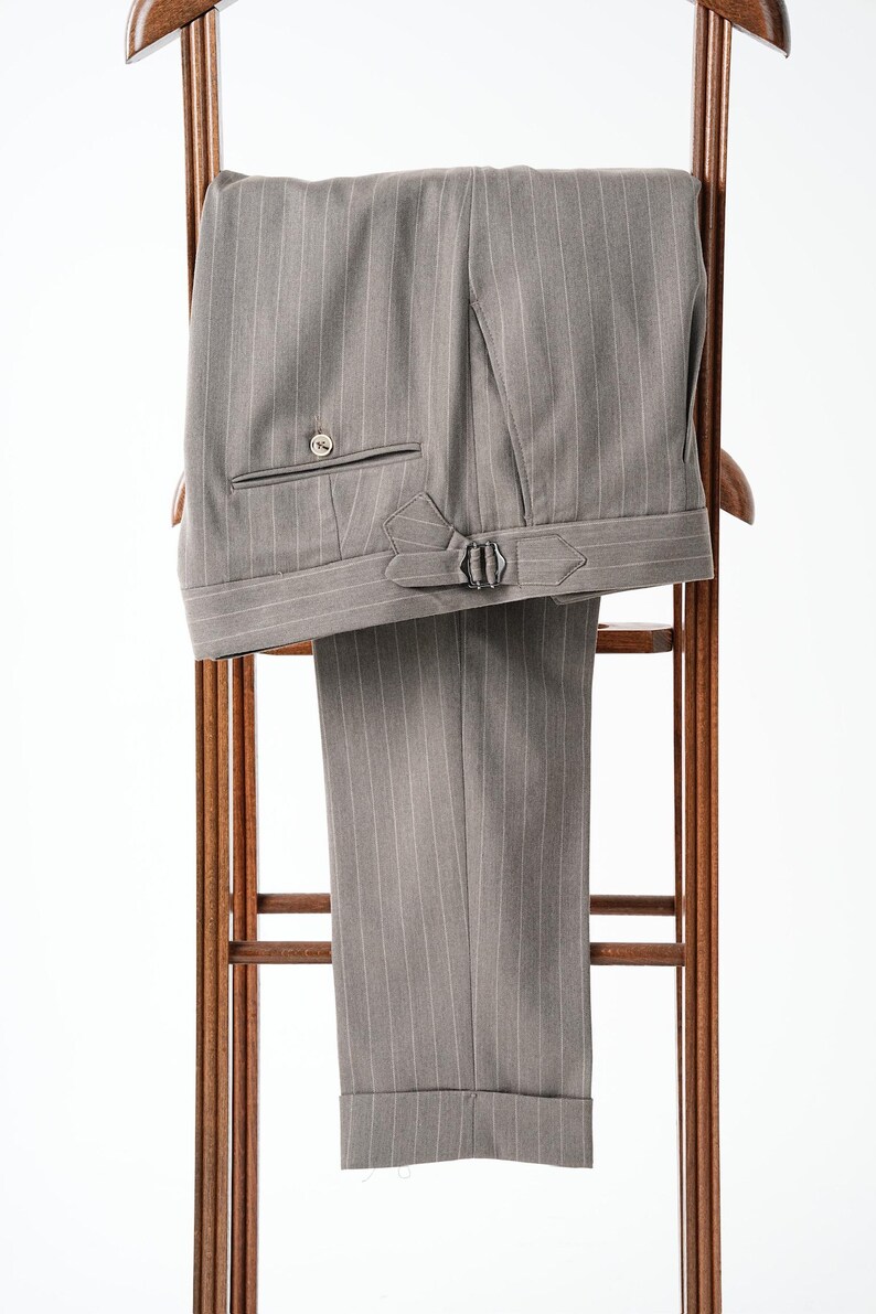 Puede incluir: Pantalones grises de raya diplom&aacute;tica con bolsillo abotonado y trabillas laterales ajustables, colgados en una percha de madera. Los pantalones tienen dobladillo con vuelta y un dise&ntilde;o cl&aacute;sico a medida, adecuado para vestimenta formal.