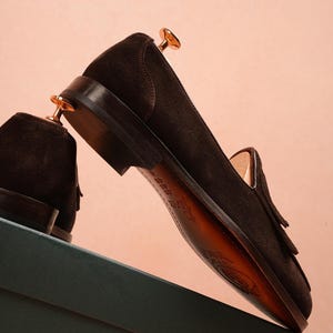 May include: A pair of dark brown suede loafers with leather soles, displayed on a green box against a pink backdrop. The shoes feature a classic tassel design. The shoes are new and unworn.