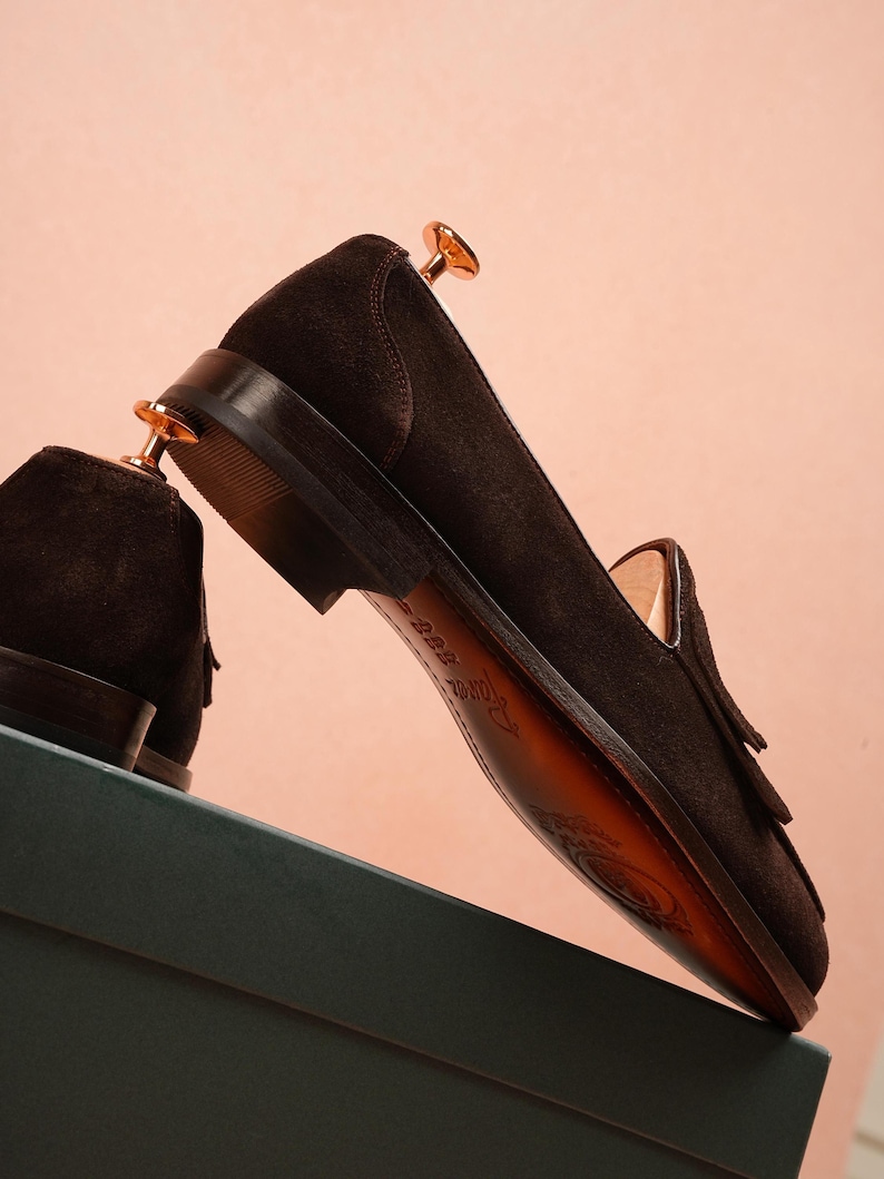 May include: A pair of dark brown suede loafers with leather soles, displayed on a green box against a pink backdrop. The shoes feature a classic tassel design. The shoes are new and unworn.