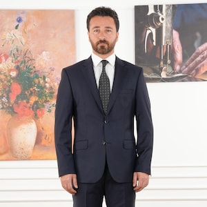 May include: A man in a navy blue suit with a black patterned tie stands in front of two paintings. The painting on the left depicts a vase of flowers, while the painting on the right shows hands working on a sewing machine.