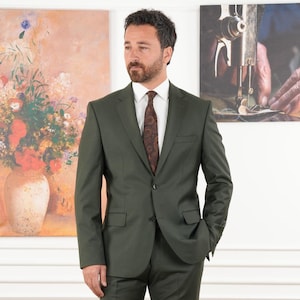 May include: A dark green suit with a white dress shirt and a patterned brown tie. The suit has a two-button closure and a notched lapel. The man is standing in front of a painting of flowers in a vase.