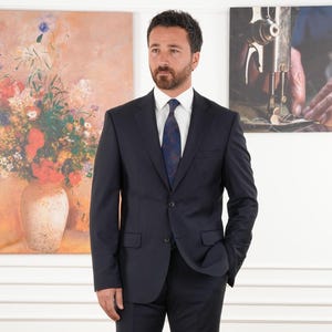 May include: A man in a navy blue suit, white shirt, and patterned tie stands in front of two framed paintings. The paintings depict a floral still life and a close-up of a sewing machine. The suit has a two-button closure.