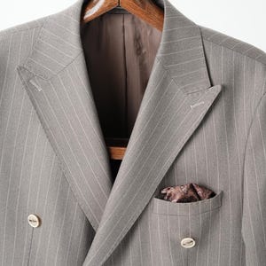 Puede incluir: Chaqueta de traje gris de doble botonadura con finas rayas blancas, colgada de una percha de madera. La chaqueta tiene solapa con muescas, dos filas de botones y un pa&ntilde;uelo de bolsillo. El forro es marr&oacute;n oscuro.