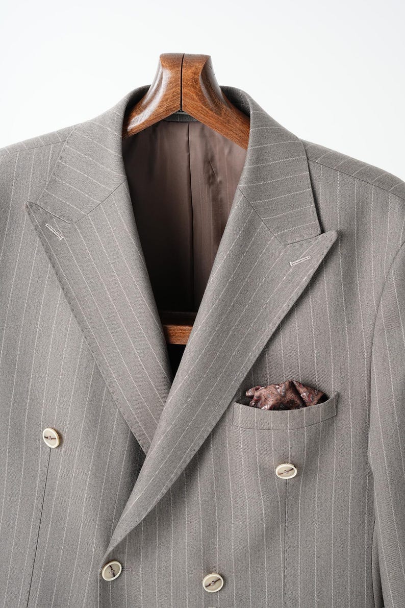 Puede incluir: Chaqueta de traje gris de doble botonadura con finas rayas blancas, colgada de una percha de madera. La chaqueta tiene solapa con muescas, dos filas de botones y un pa&ntilde;uelo de bolsillo. El forro es marr&oacute;n oscuro.