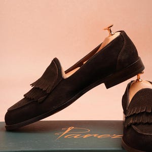May include: Dark brown suede loafers with tassel detailing, presented with wooden shoe trees. The shoes are set against a soft pink backdrop. The brand name "Farez" is visible on the box, which is dark green.