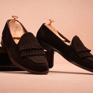 May include: A pair of dark brown suede loafers with fringe detailing. The shoes are displayed with wooden shoe trees inside, set against a soft pink background. The loafers have a classic design, suitable for various occasions.