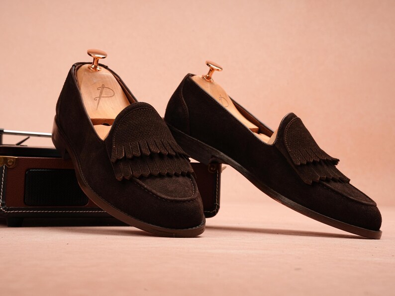 May include: A pair of dark brown suede loafers with fringe detailing. The shoes are displayed with wooden shoe trees inside, set against a soft pink background. The loafers have a classic design, suitable for various occasions.