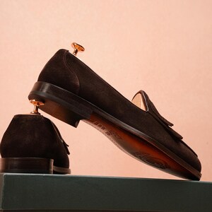 May include: Dark brown suede loafers with tassel detailing. The shoes are displayed on a green box against a pink background. The soles are a lighter brown colour. Shoe trees are inserted in the shoes.