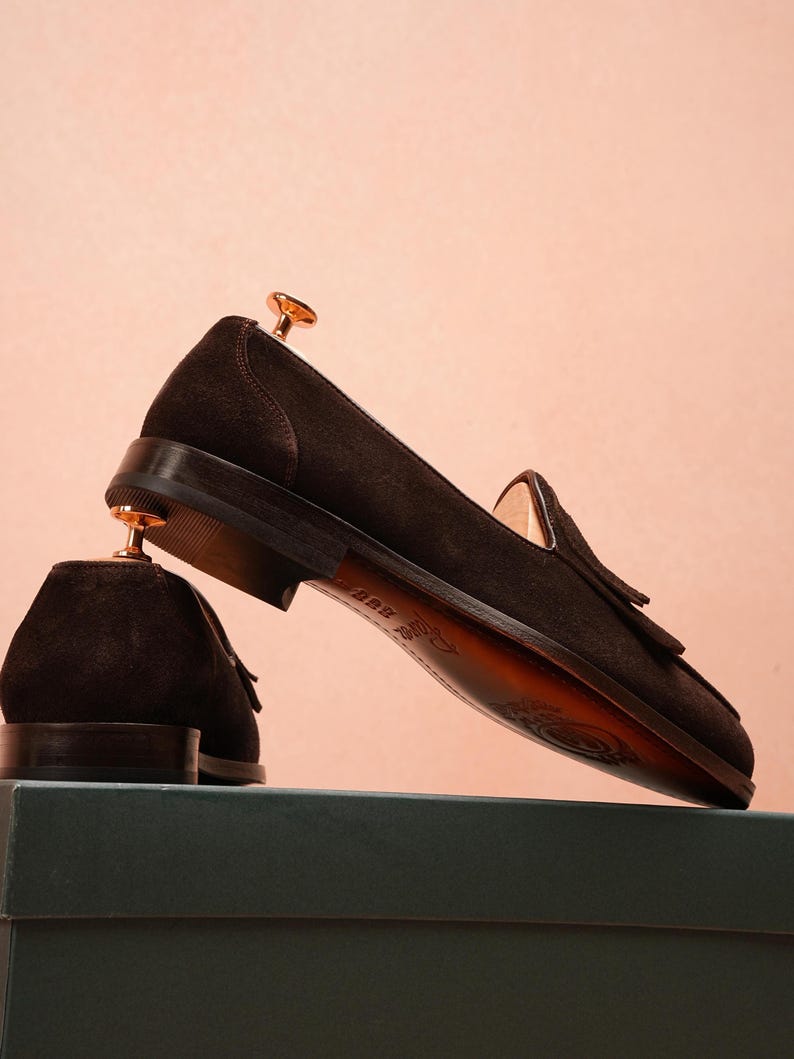 May include: Dark brown suede loafers with tassel detailing. The shoes are displayed on a green box against a pink background. The soles are a lighter brown colour. Shoe trees are inserted in the shoes.