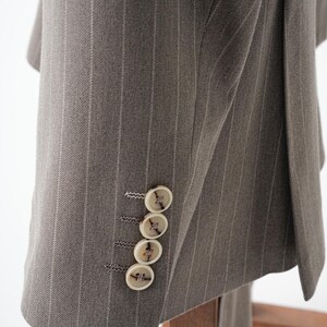 Puede incluir: Primer plano de una chaqueta de traje gris con finas rayas. La chaqueta tiene cuatro botones color crema en la manga. La tela tiene una textura sutil y las rayas son de un gris m&aacute;s claro.