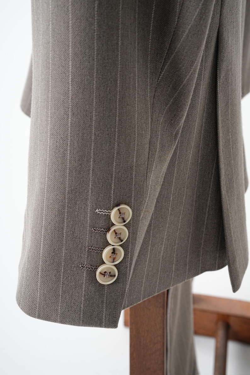 Puede incluir: Primer plano de una chaqueta de traje gris con finas rayas. La chaqueta tiene cuatro botones color crema en la manga. La tela tiene una textura sutil y las rayas son de un gris m&aacute;s claro.