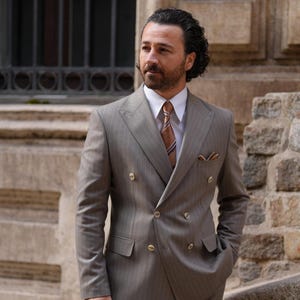 Puede incluir: Un traje cruzado marr&oacute;n claro con un sutil patr&oacute;n de rayas finas. El traje tiene botones dorados, una camisa blanca y una corbata estampada. Un pa&ntilde;uelo de bolsillo a&ntilde;ade un toque de detalle. El hombre est&aacute; afuera.