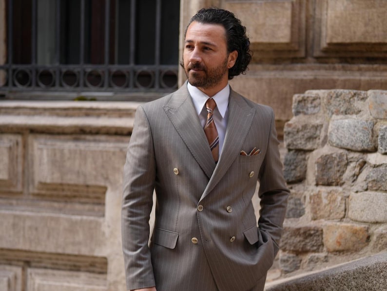 Puede incluir: Un traje cruzado marr&oacute;n claro con un sutil patr&oacute;n de rayas finas. El traje tiene botones dorados, una camisa blanca y una corbata estampada. Un pa&ntilde;uelo de bolsillo a&ntilde;ade un toque de detalle. El hombre est&aacute; afuera.